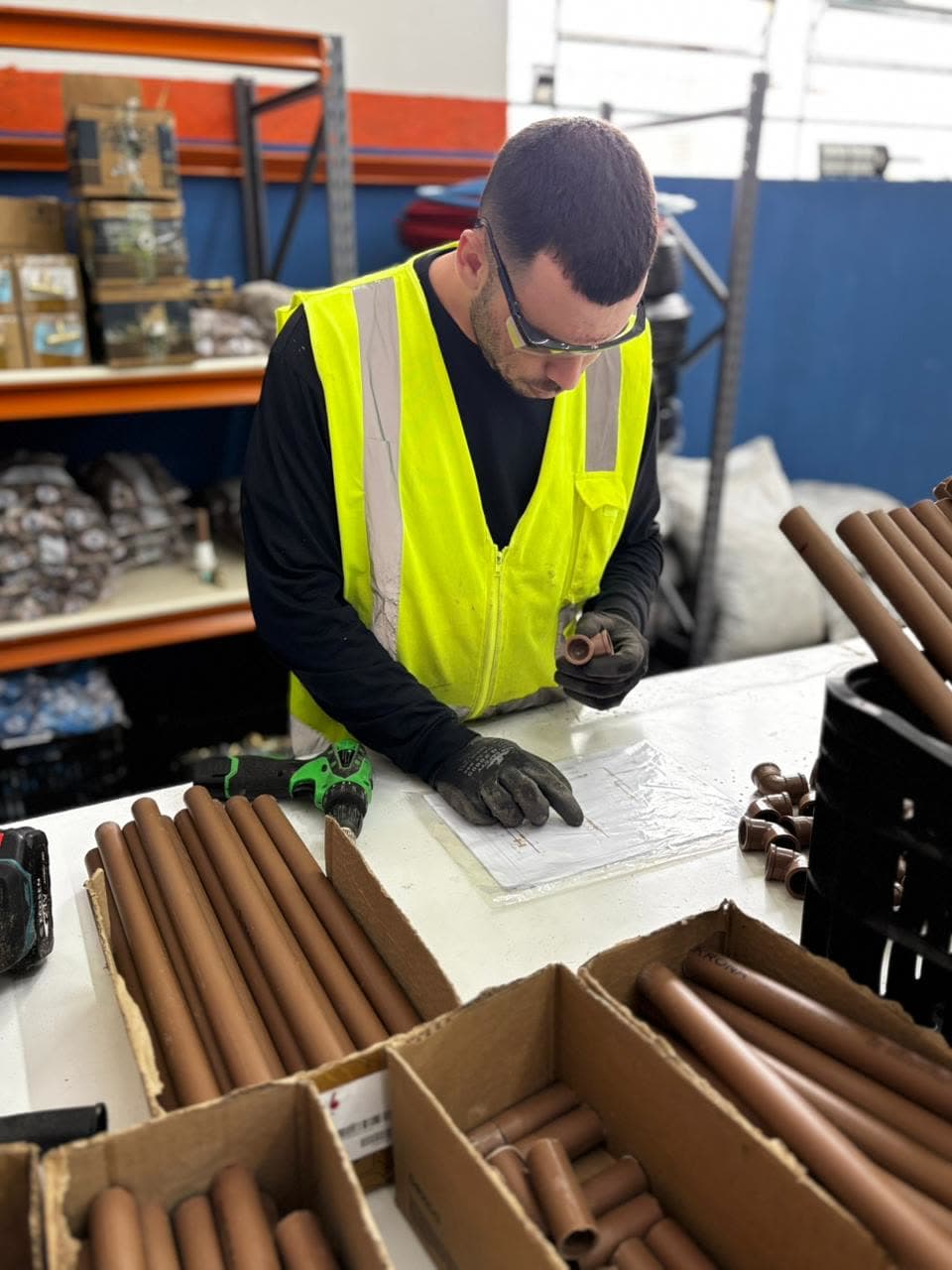 Montador da Polvolt de colete refletivo e óculos de proteção conferindo o projeto plastificado na bancada, entre tubos de PPR cortados e conexões